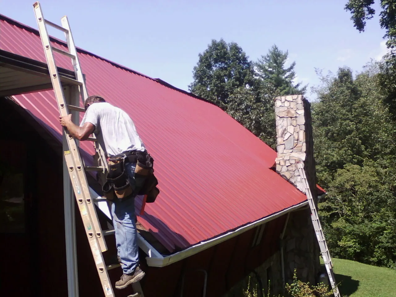 Roofer on a ladder beside a red metal roof and stone chimney — Soffit & Fascia Repair by Roofing Brothers in Joshua